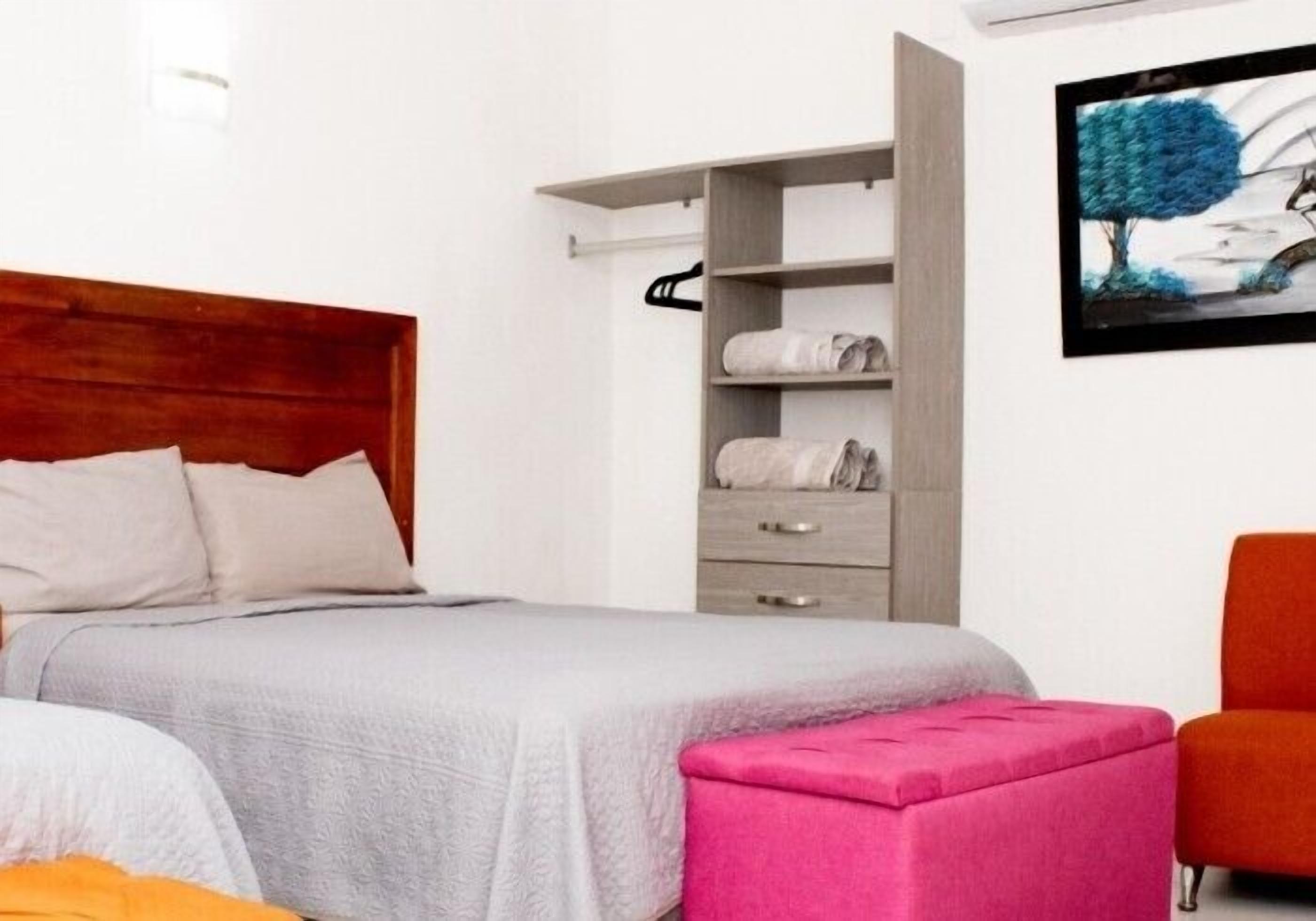Une chambre à coucher avec un lit, une tête de lit en bois, une armoire, un pouf rose et un tableau encadré accroché au mur.