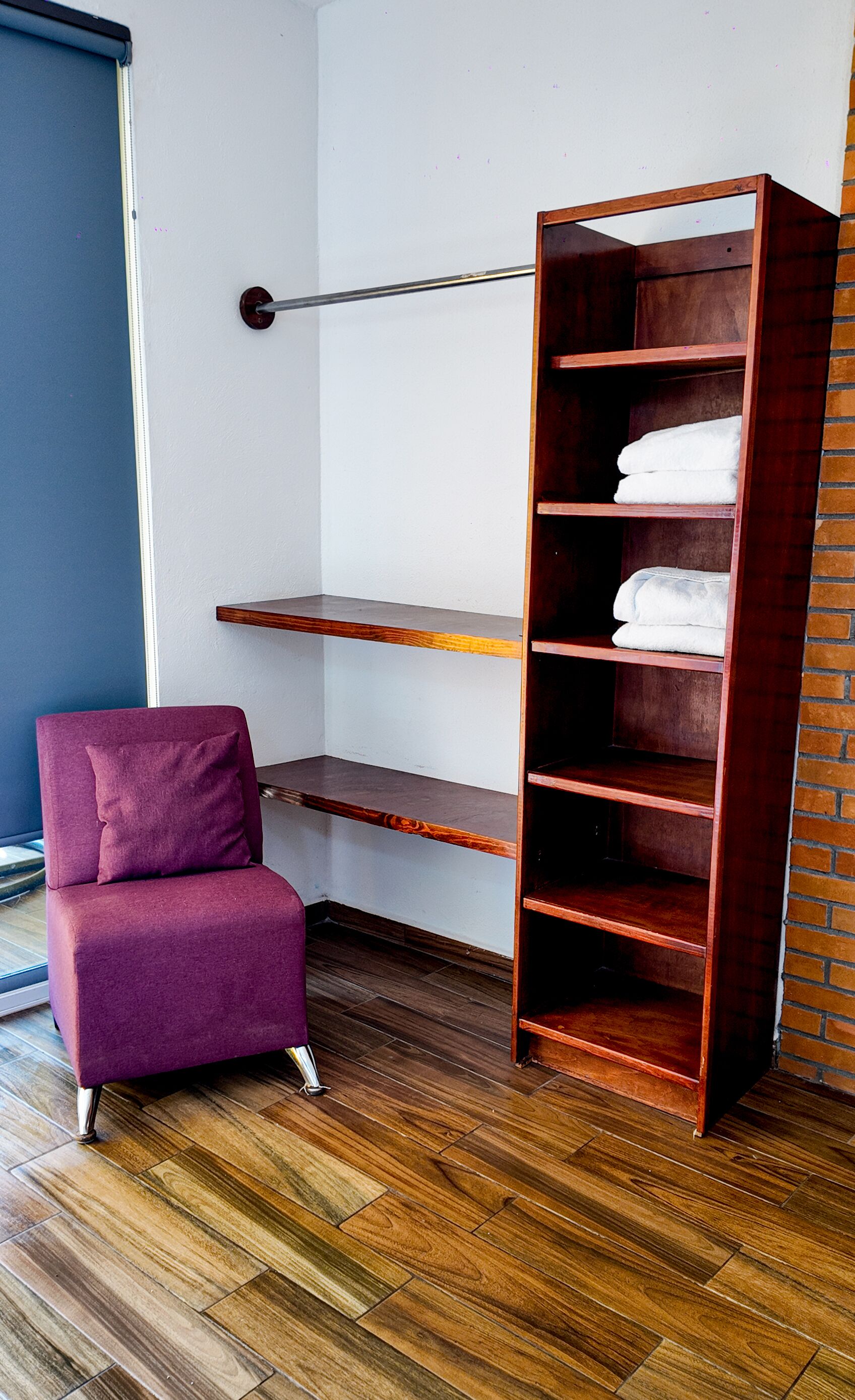 A room with a wooden floor, a purple armchair, a tall wooden bookshelf, and a brick wall.