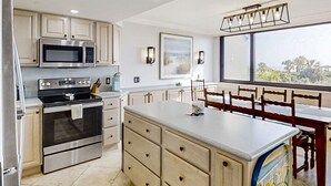 A modern kitchen with stainless steel appliances, a dining area with wooden chairs, and a large window offering a view. - Spacious condo with beach access, balcony, AC & outdoor pool (Fernandina Beach)