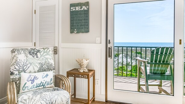 A room with a view of the sea, a chair with a cushion, a small table with a decorative item, and a sign with motivational text.