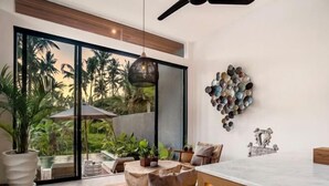 A modern living room with a large sliding glass door leading to a tropical garden, a dining area with a marble table, and a wall-mounted mosaic art piece. - Villa Ornella by BaliSuperHost (Bali)
