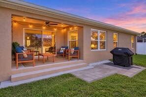 Fachada de una vivienda con porche cubierto, mobiliario exterior y una parrilla de barbacoa.