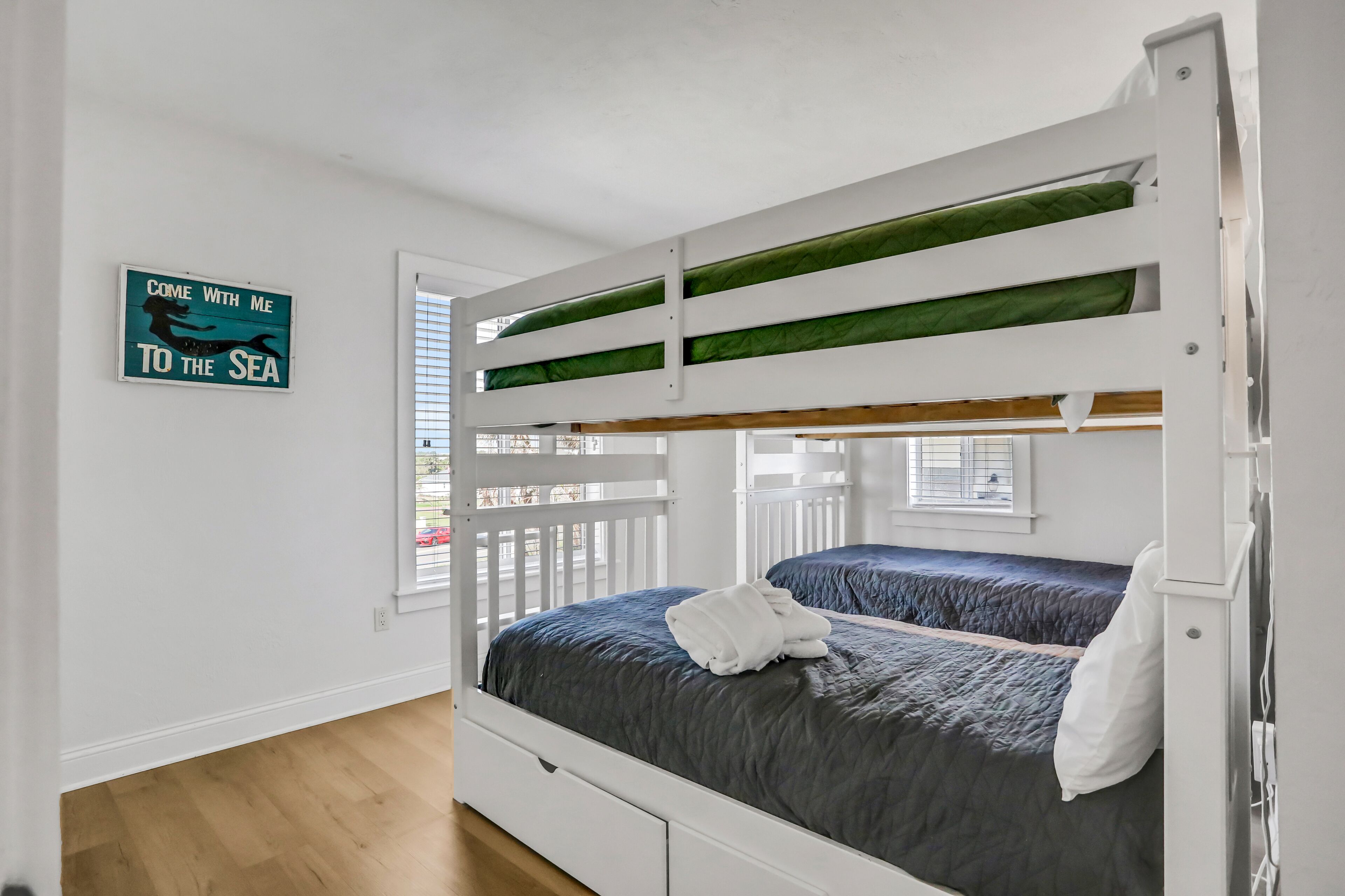 Quarto com beliches, placa na parede, piso de madeira e janela com persianas.