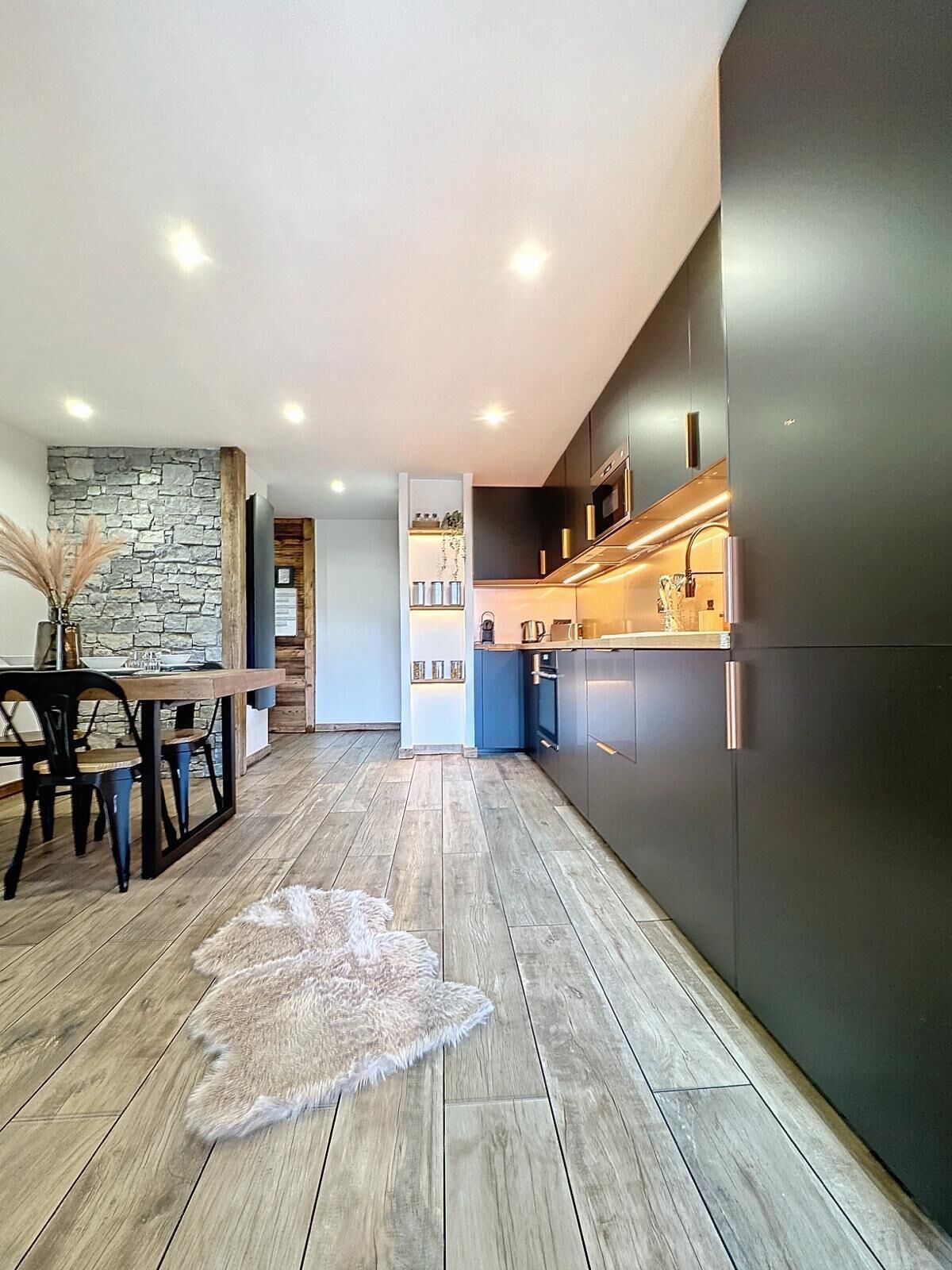 Une cuisine moderne avec des meubles de couleur sombre, une table à manger en bois et un mur décoratif en pierre.