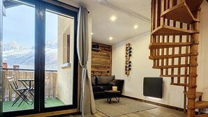 Amplia sala de estar con piso de madera, un sofá, una mesa de centro y una chimenea. Una gran puerta corrediza de vidrio da a un balcón con vistas a montañas.