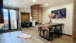 Una sala de estar moderna con piso de madera, un comedor con mesa y sillas de madera, un televisor grande montado en la pared y vistas al exterior a través de una puerta corrediza de vidrio.