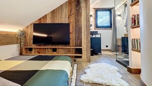 Un dormitorio moderno con techo de madera, un televisor de pantalla plana grande, una cama con muchos almohadones, un tapete de piel blanca y una estantería.