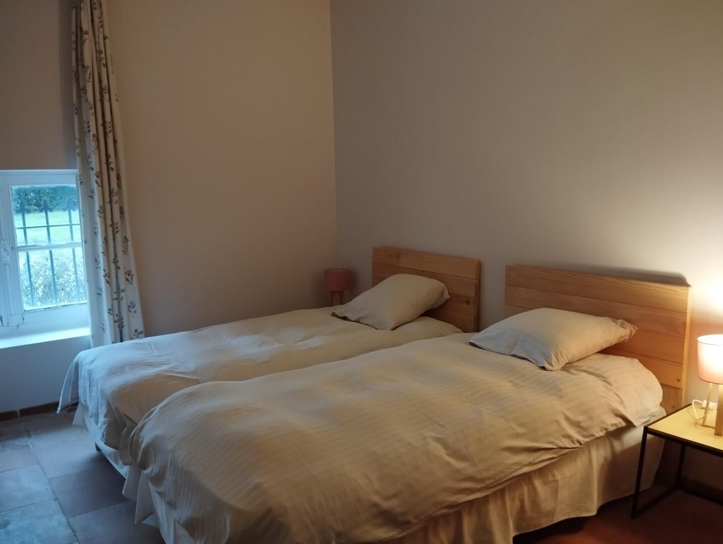 A room with two single beds, a window with curtains, a bedside table with a lamp, and a view of greenery outside.
