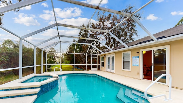 A covered outdoor swimming pool with a glass enclosure, a hot tub, and a poolside area with a door leading to a building.