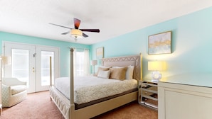 A bedroom with a four-poster bed, a ceiling fan, a bedside table with a lamp, and a door with a view.