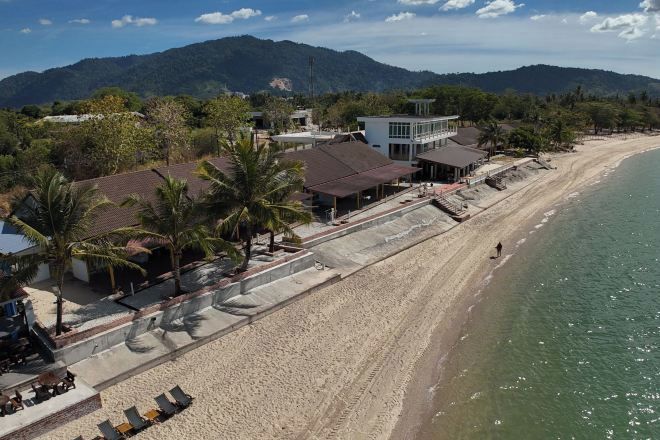 A beachside property with a long sandy beach, a few palm trees, and a mountain backdrop.