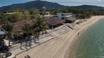 A beachside property with a long sandy beach, a few palm trees, and a mountain backdrop.