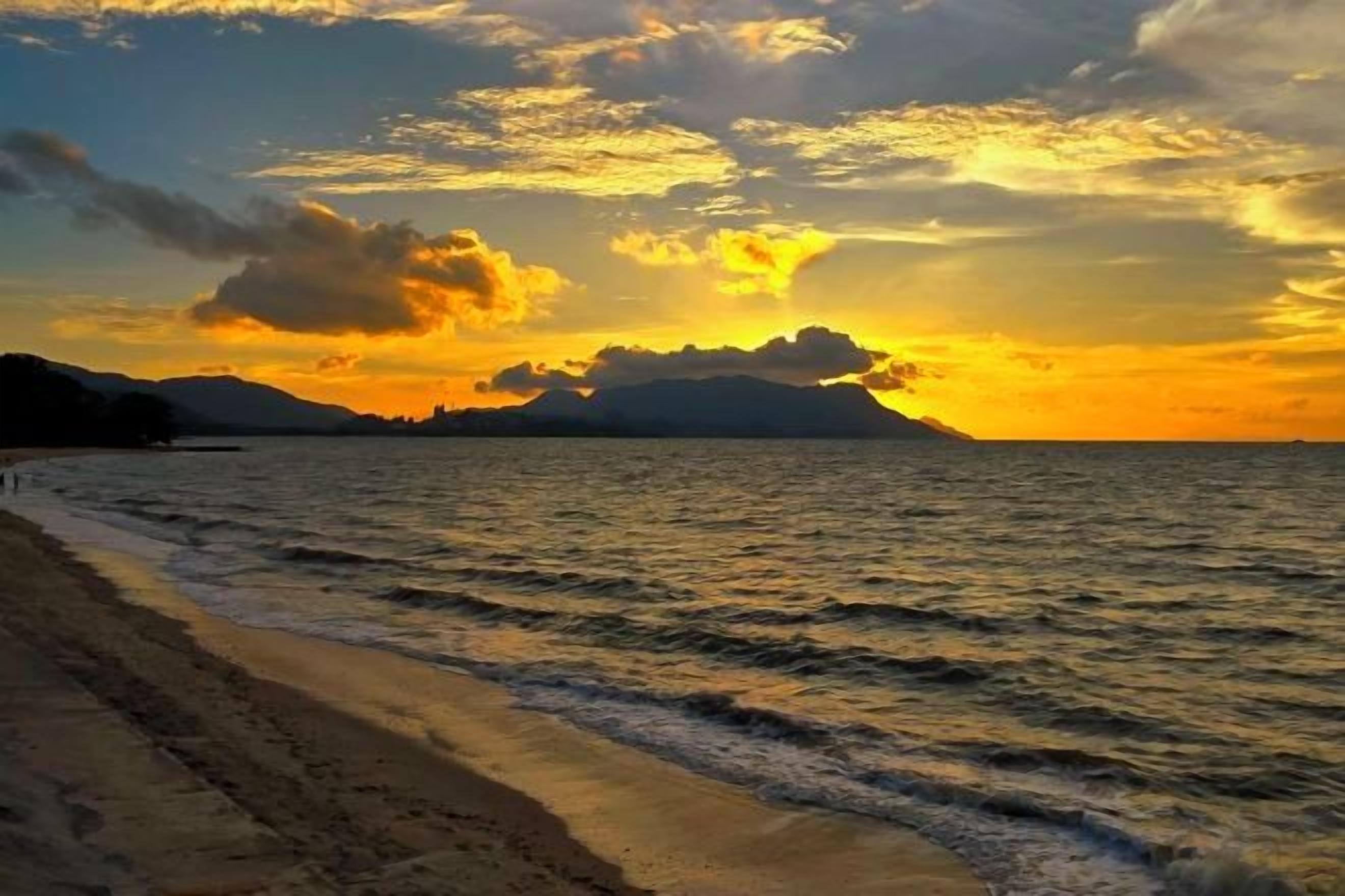 Uma praia com ondas, um pôr do sol e montanhas ao longe.