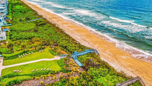 An aerial view of a coastal hotel with a beach, a golf course, and a blue staircase leading to the beach. - Coastal Gem on the Golf Course w/ Shared Pool, & Beach Access - Dog-Friendly (Palm Coast)