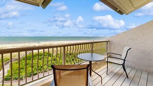 A balcony with a view of the beach, a table, and two chairs.