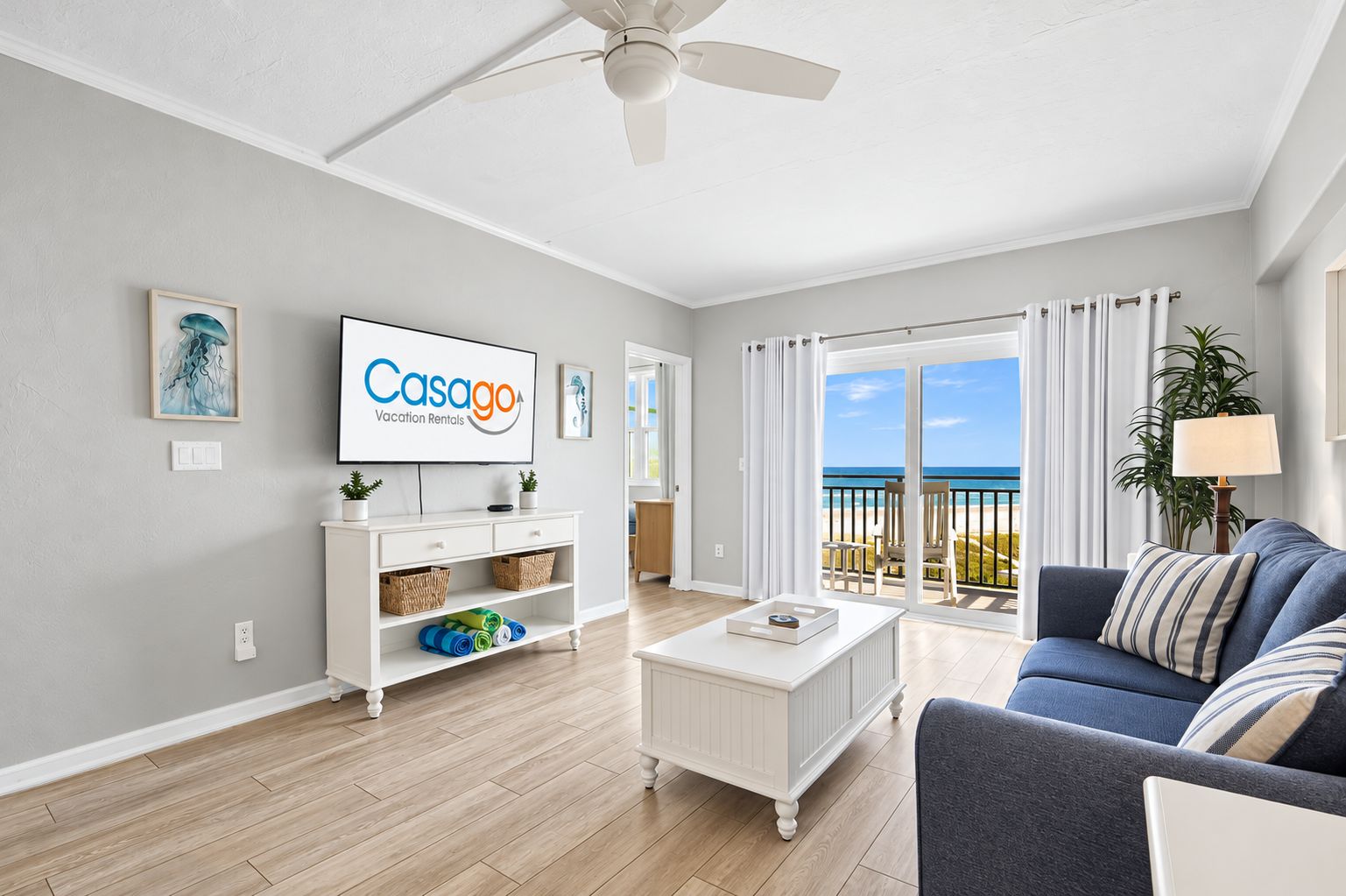 A modern living room with a flat-screen TV, a white coffee table, a blue sofa, and a view of the beach through large windows.
