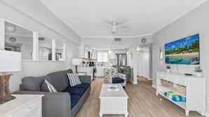 Una sala de estar moderna con un sofá, una mesa de centro y un televisor. Se observa una cocina al fondo con una zona de comedor.