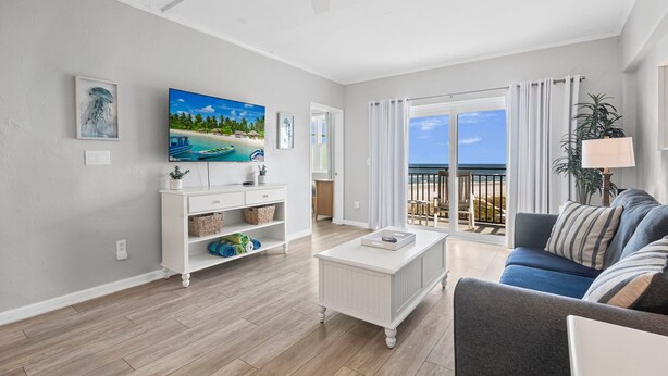 Una sala de estar con televisor, muebles blancos, un balcón con vistas y un ventilador de techo.