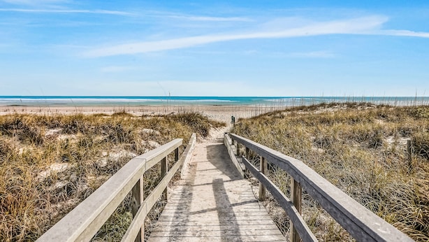 Une promenade en bois qui mène à une plage bordée de dunes, sous un ciel dégagé.
