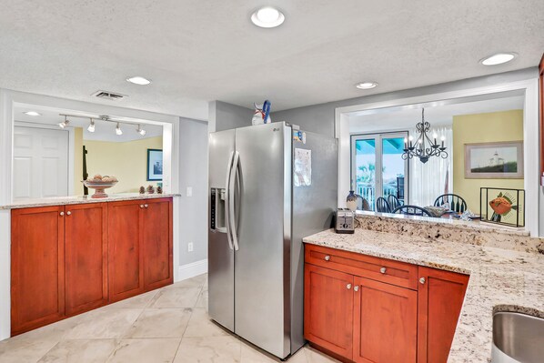 A modern kitchen with wooden cabinets, a stainless steel refrigerator, granite countertops, and recessed ceiling lights. - Oceanfront condo with direct beach access, pool, balcony, grilling area, & AC (Fernandina Beach)