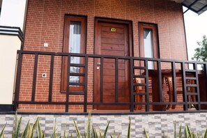 A brick building with a wooden door and windows, a metal railing, and a checkered pattern at the base. - Sunny Point Penida (Bali)