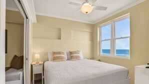 A bedroom with a bed, bedside tables, a nightstand, a ceiling fan, and a window with a view of the ocean. - Oceanfront condo with pool, amazing view, balcony & beach access (Jacksonville Beach)