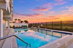 A swimming pool with a sunset view, a fence, and palm trees. - Oceanfront condo with pool, amazing view, balcony & beach access (Jacksonville Beach)