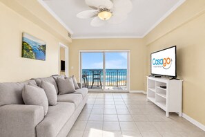 A living room with a sofa, a television, and a balcony with a view of the beach.