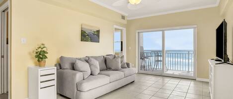 A living room with a sofa, a small white cabinet, a ceiling fan, a large window with a view, and a balcony.