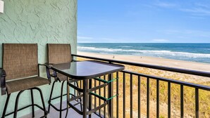 A balcony with two chairs and a table, overlooking a beach with waves and a clear sky. - Oceanfront condo with pool, amazing view, balcony & beach access (Jacksonville Beach)