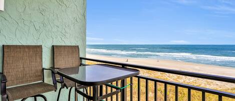 A balcony with two chairs and a table, overlooking a beach with waves and a clear sky.