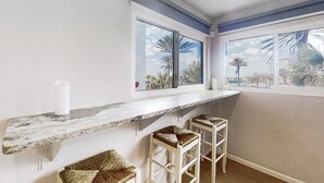 Habitación con encimera de mármol, dos taburetes de mimbre y vistas a palmeras a través de amplios ventanales.