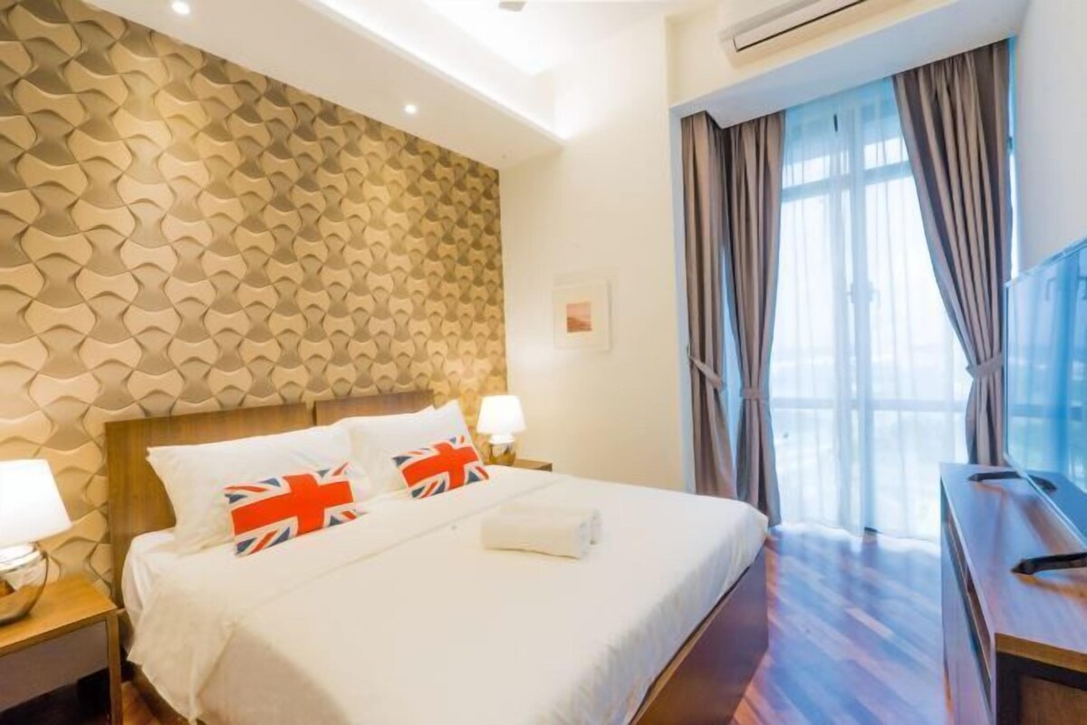 A hotel room with a patterned wall, a bed with pillows, a TV, and a window with curtains.