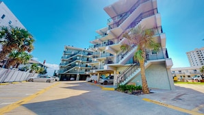 A multi-story residential building with balconies, a parking area, and palm trees.