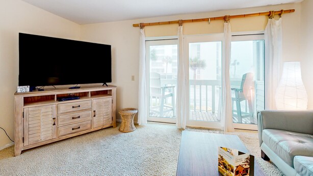 Salón con televisor de pantalla plana, mueble de entretenimiento de madera, sofá gris y balcón con vista a palmeras.