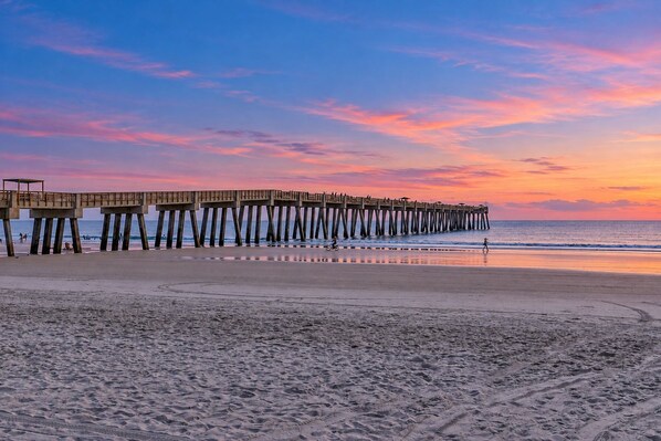 A beach with a wooden pier extending into the ocean, sandy beach, and a sunset sky.