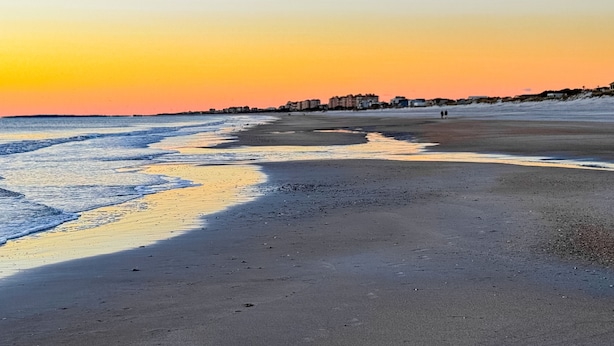 En strand med solnedgang, rolige vand og bygninger i baggrunden.