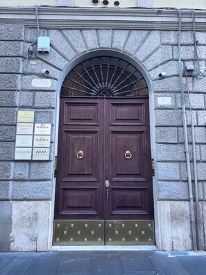 A double wooden door with a metal grill, a stone wall with a sign, and a metal pipe running along the wall.