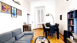 A living room with a grey sofa, a coffee table with a bowl, a dining table with chairs, a bookshelf, and a window with curtains. - Beauty House in Cola di Rienzo• 4 Bedrooms - 2 Baths - St. Peter's, Central Rome (Roma)
