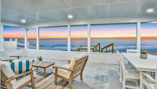 Área de estar coberta ao ar livre com vista para o oceano ao pôr do sol, com móveis de madeira e uma mesa branca.