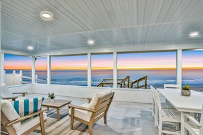 Un espace extérieur abrité, aménagé avec des meubles en bois et une table blanche, offrant une vue sur l’océan au coucher du soleil.
