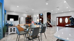 A spacious living area with a glass dining table, comfortable seating, a staircase, and a large wooden door.