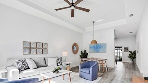 Een moderne woonkamer met een witte bank, een blauwe fauteuil en een salontafel. Er is een eethoek met een tafel en stoelen, en een plafondventilator.