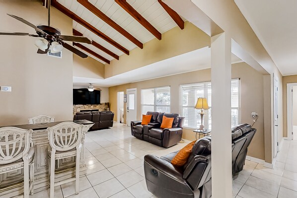 A spacious living room with a high ceiling, a dining area with white chairs, and a black leather sofa with orange cushions.