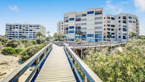 Eine Holzpromenade, die zu einem Hotel am Strand mit mehreren Etagen und Balkonen führt.