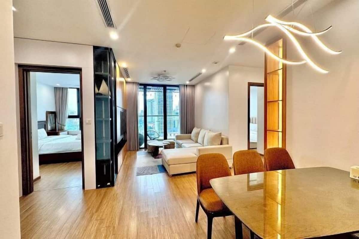 Quarto de hotel moderno com piso de madeira, área de refeições com mesa e cadeiras, um sofá e vista da cidade através de grandes janelas.