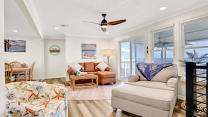 A living room with a sofa, armchair, and coffee table. There is a ceiling fan, a painting, and a window with blinds.