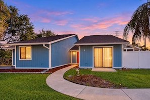 A two-story residential house with a blue exterior, a front yard with a curved pathway, and a white fence. - Water-view dog-friendly home with outdoor space & entertainment options (Palm Coast)