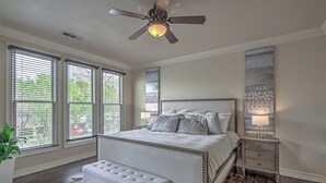 A bedroom with a large bed, a bench, a nightstand, a ceiling fan, and a window with blinds.