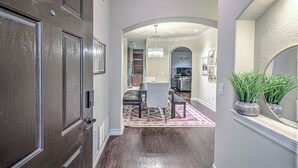 A hallway with a dark wooden door, a rug, a dining area with a chandelier, and a mirror with plants.